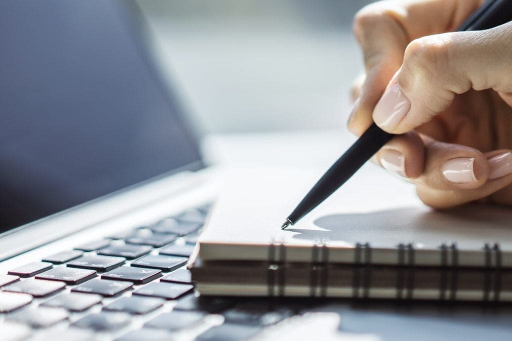 Hand writing notes in a notebook beside a laptop keyboard